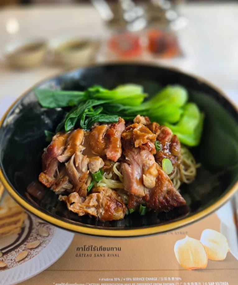 黒いボウルにきれいに盛り付けられた、たっぷりの青菜とジューシーなローストポークがのった卵麺（バミー）料理。